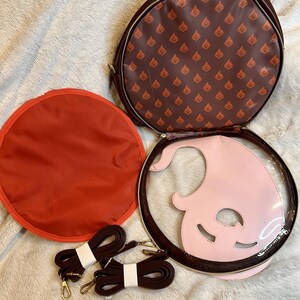 May include: A brown round bag with a pink and clear plastic insert. The bag has a brown strap and a red lining. The bag has a repeating pattern of a flame design on the outside.