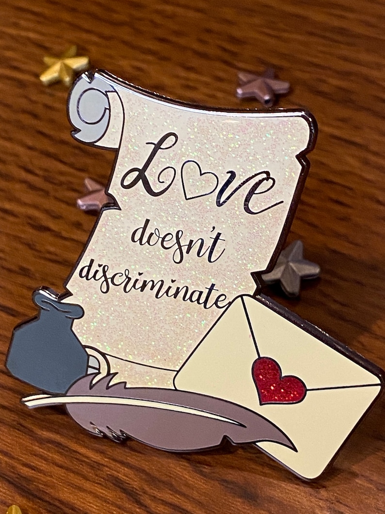 Pode incluir: Um pergaminho branco com glitter e o texto "O amor n&atilde;o discrimina" com um cora&ccedil;&atilde;o vermelho, uma pena cinza, um tinteiro cinza e um envelope branco com um cora&ccedil;&atilde;o vermelho.