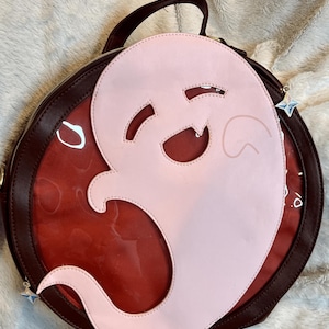 May include: A round, burgundy leather bag with a clear plastic window. The bag has a pink ghost cutout with a smiling face. The bag has a top handle and a detachable star charm.