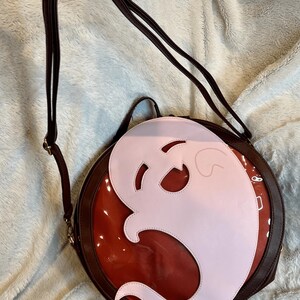 May include: A round, brown leather purse with a clear plastic window. The window shows a pink ghost shape.