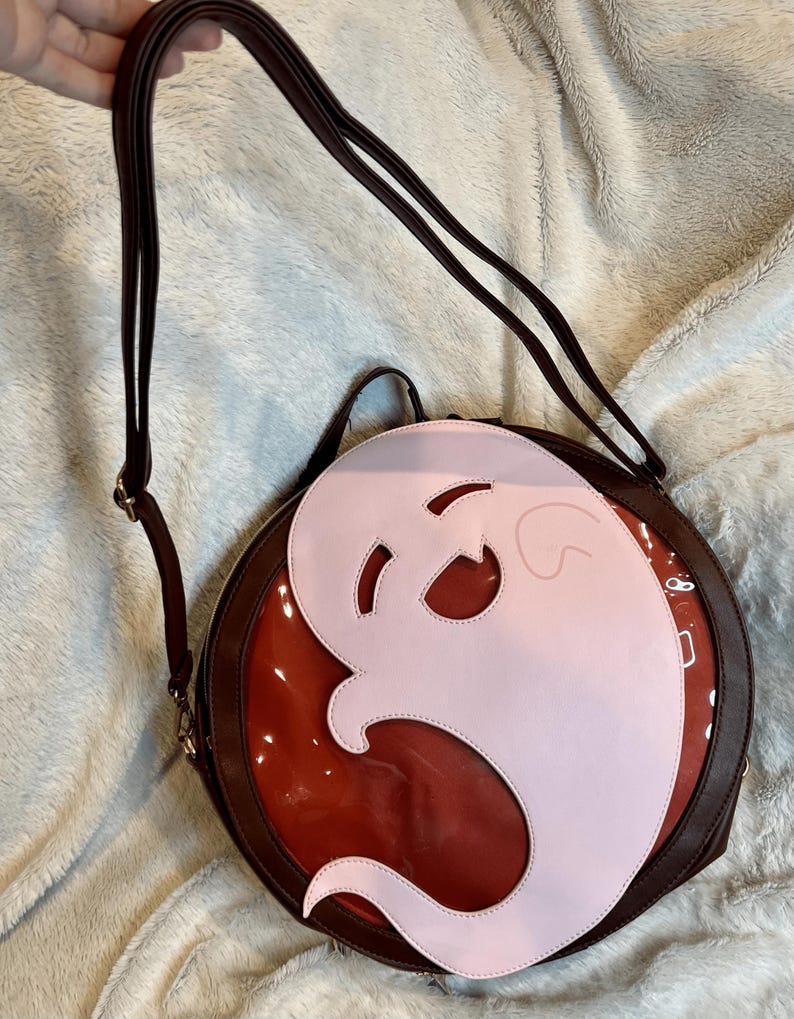 May include: A round, brown leather purse with a clear plastic window. The window shows a pink ghost shape.