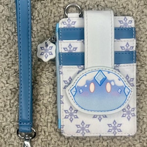 May include: A white and blue wallet with a matching wrist strap. The wallet features a snowflake pattern and a cartoon character design. The wrist strap is light blue and has a silver clasp. The wallet has a zipper and multiple card slots.