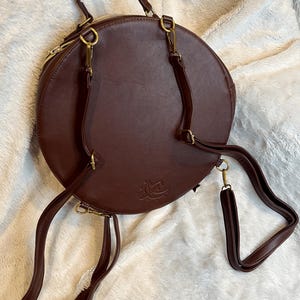 May include: A brown leather round backpack with gold hardware and adjustable straps. The bag has a zipper closure and a small logo on the front.