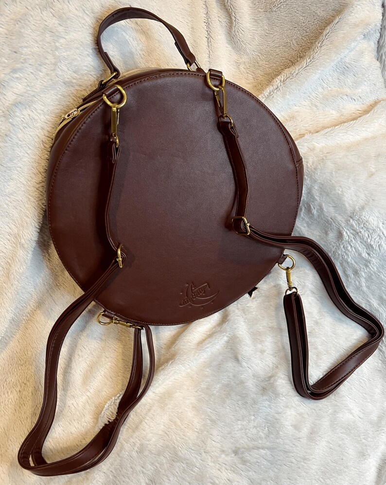 May include: A brown leather round backpack with gold hardware and adjustable straps. The bag has a zipper closure and a small logo on the front.