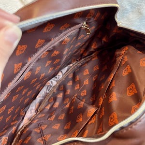 May include: A brown leather backpack with a zippered pocket and a patterned lining. The lining features a repeating pattern of a stylized orange bear head.