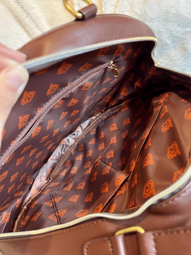 May include: A brown leather backpack with a zippered pocket and a patterned lining. The lining features a repeating pattern of a stylized orange bear head.