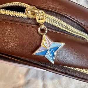 May include: A brown leather bag with a gold zipper and a star-shaped charm. The charm is made of metal and has a blue, pink, and yellow design.