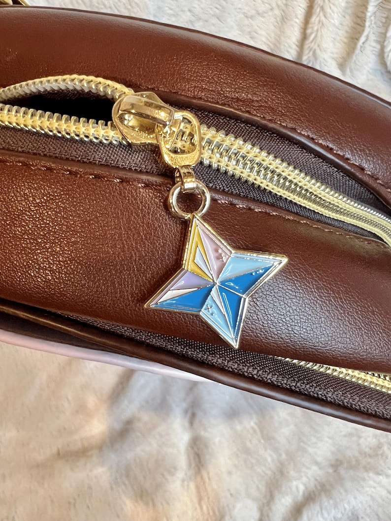 May include: A brown leather bag with a gold zipper and a star-shaped charm. The charm is made of metal and has a blue, pink, and yellow design.