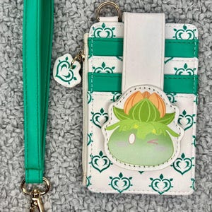 May include: A white and green cardholder with a matching green wrist strap. The cardholder features a cartoon character design and a zipper closure. The wrist strap has a gold-colored clasp.