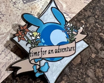 Op de afbeelding: Een blauwe emaille pin met een witte achtergrond en een blauw en wit swirl-ontwerp. De pin heeft een banner met de tekst "time for an adventure" en een bloemmotief met rode, witte en blauwe bloemen.