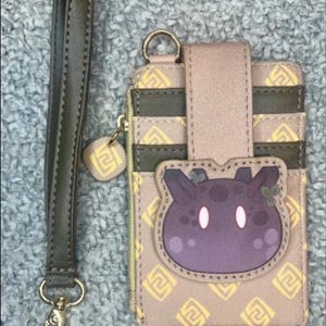 May include: A beige and olive green cardholder with a wrist strap. The cardholder features a purple cartoon character design and a gold zipper. The wrist strap is olive green with a gold clasp. The cardholder has multiple card slots.