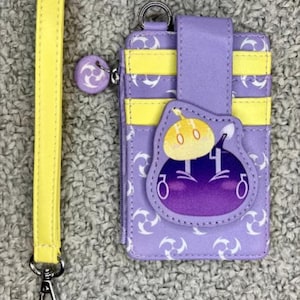 May include: A purple cardholder with yellow accents and a matching yellow wrist strap. The cardholder features a character design and a zipper closure. The wrist strap has a silver clasp. The cardholder is approximately 4 inches tall.