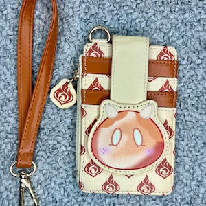 May include: A cream-colored cardholder with a cartoon character design, featuring brown accents and a matching wrist strap. The cardholder has a zipper and a metal clasp. The design includes a flame symbol.