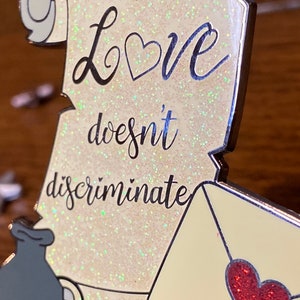 Pode incluir: Um pergaminho branco com glitter e o texto "Love doesn't discriminate" em preto. Um cora&ccedil;&atilde;o vermelho est&aacute; em um envelope amarelo no canto inferior direito.