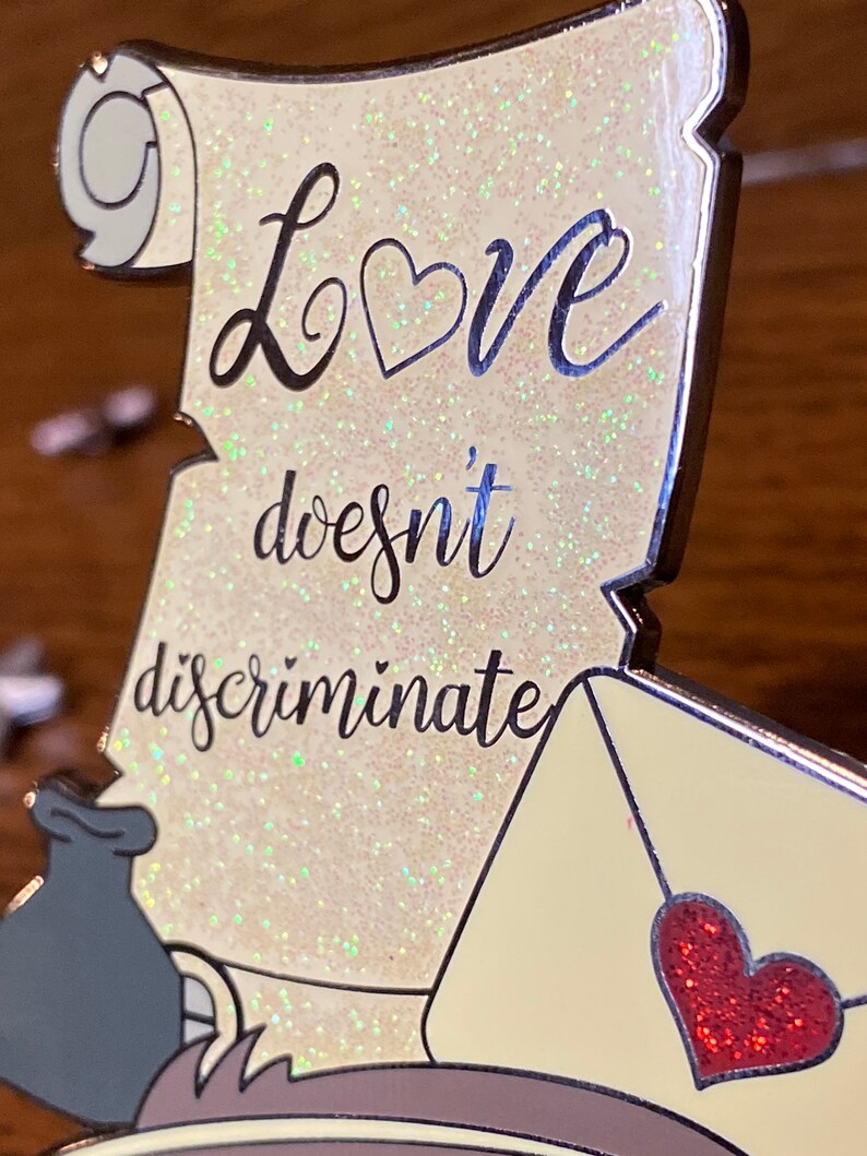 Pode incluir: Um pergaminho branco com glitter e o texto "Love doesn't discriminate" em preto. Um cora&ccedil;&atilde;o vermelho est&aacute; em um envelope amarelo no canto inferior direito.
