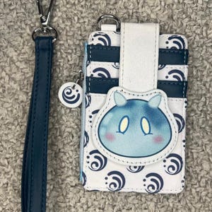 May include: A white and blue card holder with a blue and white swirl pattern. The card holder has a blue character with white eyes and pink cheeks on the front. The card holder has a detachable strap.