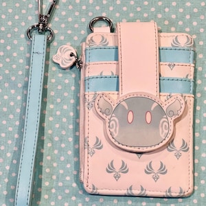 May include: A light pink and blue cardholder with a matching wrist strap. The cardholder features a cartoon character design and a pattern of stylized wings. The wrist strap and cardholder have silver hardware.