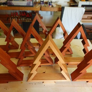 May include: A set of eight wooden Christmas tree-shaped shelves, some stained a darker brown and others a lighter natural wood color. The shelves are arranged in a staggered pattern on a light wood surface.