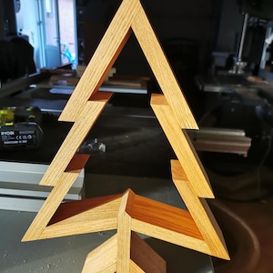 May include: A wooden Christmas tree ornament crafted from light brown wood. The tree is constructed from multiple pieces of wood, joined together to form a three-dimensional shape. The tree has a simple design and is a great way to add a touch of rustic charm to your festive decorations.