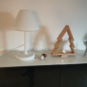 May include: A white table lamp with a white shade sits on a white surface next to a wooden Christmas tree ornament. A small candle is lit inside the ornament.