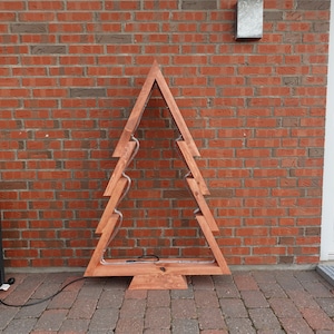 Puede incluir: Decoración de madera en forma de árbol de Navidad con acabado marrón cálido. El árbol tiene un contorno simple con un diseño en zigzag y una tira de luz en el interior. Se encuentra sobre un patio de ladrillos contra una pared de ladrillo rojo.
