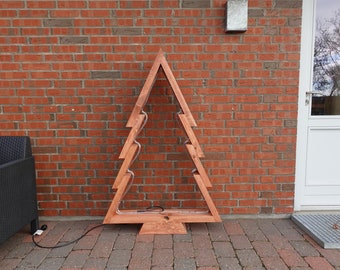 May include: A wooden Christmas tree-shaped decoration with a warm brown finish. The tree has a simple outline with a zigzag design and a light strip inside. It stands on a brick patio against a red brick wall.