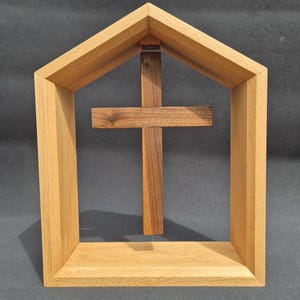 Op de afbeelding: Een lichtgekleurd houten frame in de vorm van een huis met een puntdak, met daarin een donkerhouten kruis dat in het midden hangt. Het kruis is verticaal in de open structuur geplaatst, wat een contrast vormt tussen de twee houttinten.