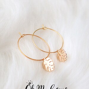 May include: A pair of gold hoop earrings featuring small, dangling monstera leaf charms. The jewelry is positioned above the text Oh My Gems DESIGN, which includes a small icon of a ring with a diamond above the letter O.