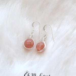 May include: Silver dangle earrings featuring pink, teardrop-shaped gemstones. The gemstones are encased in silver wire and suspended from silver ear wires. The earrings are presented on a white, fluffy background.