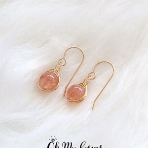 Peut inclure: Une paire de boucles d'oreilles pendantes avec des perles de pierre de soleil rondes couleur pêche, entourées de fil doré et munies de crochets dorés. Le texte Oh My Gems DESIGN est imprimé en noir sous les boucles d'oreilles.