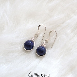 May include: A pair of silver wire wrapped earrings with dark blue gemstone drops. The earrings are on a white fluffy surface.