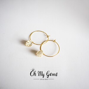 May include: A pair of gold hoop earrings featuring a small, textured monstera leaf charm attached to each hoop. The text "Oh My Gems DESIGN" is printed in black below the earrings, with a small diamond icon above the word Oh.
