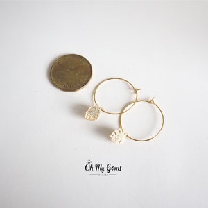 May include: A pair of gold hoop earrings featuring small, cream-colored monstera leaf charms. A Canadian one-dollar coin is placed next to the earrings for scale. The text "Oh My Gems DESIGN" is printed in black at the bottom of the image.