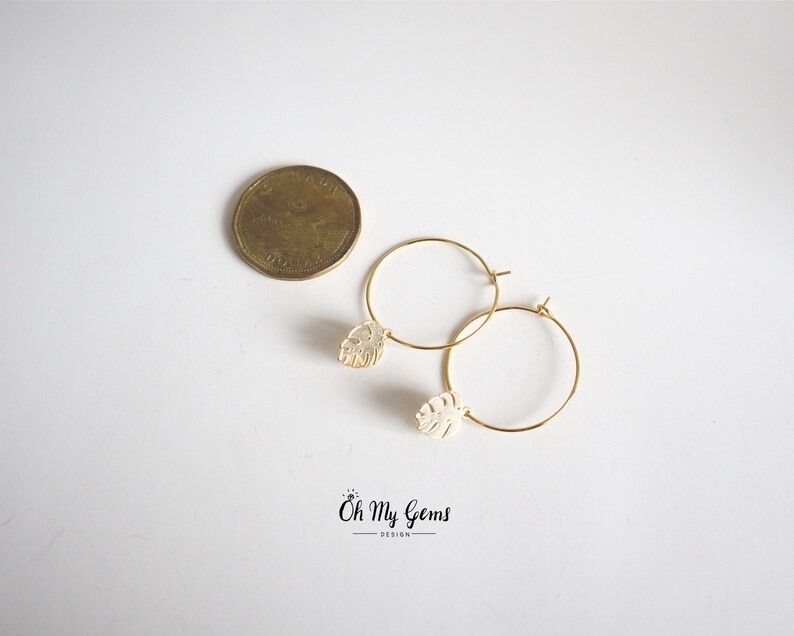 May include: A pair of gold hoop earrings featuring small, cream-colored monstera leaf charms. A Canadian one-dollar coin is placed next to the earrings for scale. The text "Oh My Gems DESIGN" is printed in black at the bottom of the image.