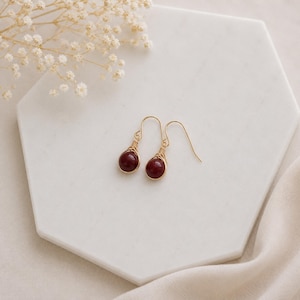 May include: A pair of drop earrings featuring round, deep red beads secured with gold-toned wire wrapping. Each earring hangs from a simple gold-toned hook, showcasing a polished finish and a minimalist design that highlights the dark red stones.