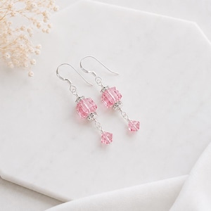 May include: A pair of drop earrings featuring faceted light pink crystal beads. Each earring consists of a larger square-cut pink crystal bead accented with silver-toned metal spacers and a smaller dangling pink crystal bead on a silver wire hook.