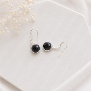 May include: A pair of dangle earrings featuring round, polished black stones encased in silver-toned wire wrapping. Each earring is suspended from a simple silver-toned hook, showcasing a clean, metallic design that secures the dark, spherical beads.