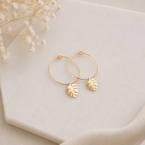 May include: A pair of gold hoop earrings featuring small, dangling monstera leaf charms. The earrings are circular with a thin wire construction and a delicate, cut-out leaf design suspended from the bottom of each hoop, showcasing a metallic finish.