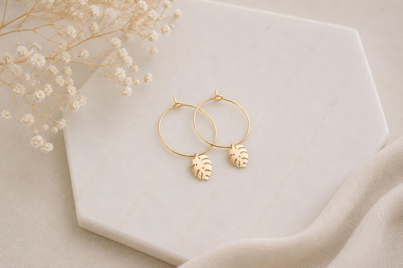 May include: A pair of gold hoop earrings featuring small, dangling monstera leaf charms. The earrings are circular with a thin wire construction and a delicate, cut-out leaf design suspended from the bottom of each hoop, showcasing a metallic finish.