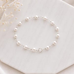 May include: A delicate bracelet featuring a series of small, round, white pearls spaced evenly along a thin, polished silver-tone metal chain. The bracelet is secured with a circular spring ring clasp, creating a continuous, circular design.