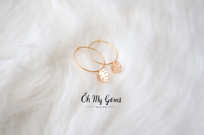 May include: A pair of gold hoop earrings featuring small, dangling monstera leaf charms. The jewelry is positioned above the text Oh My Gems DESIGN, which includes a small icon of a ring with a diamond above the letter O.