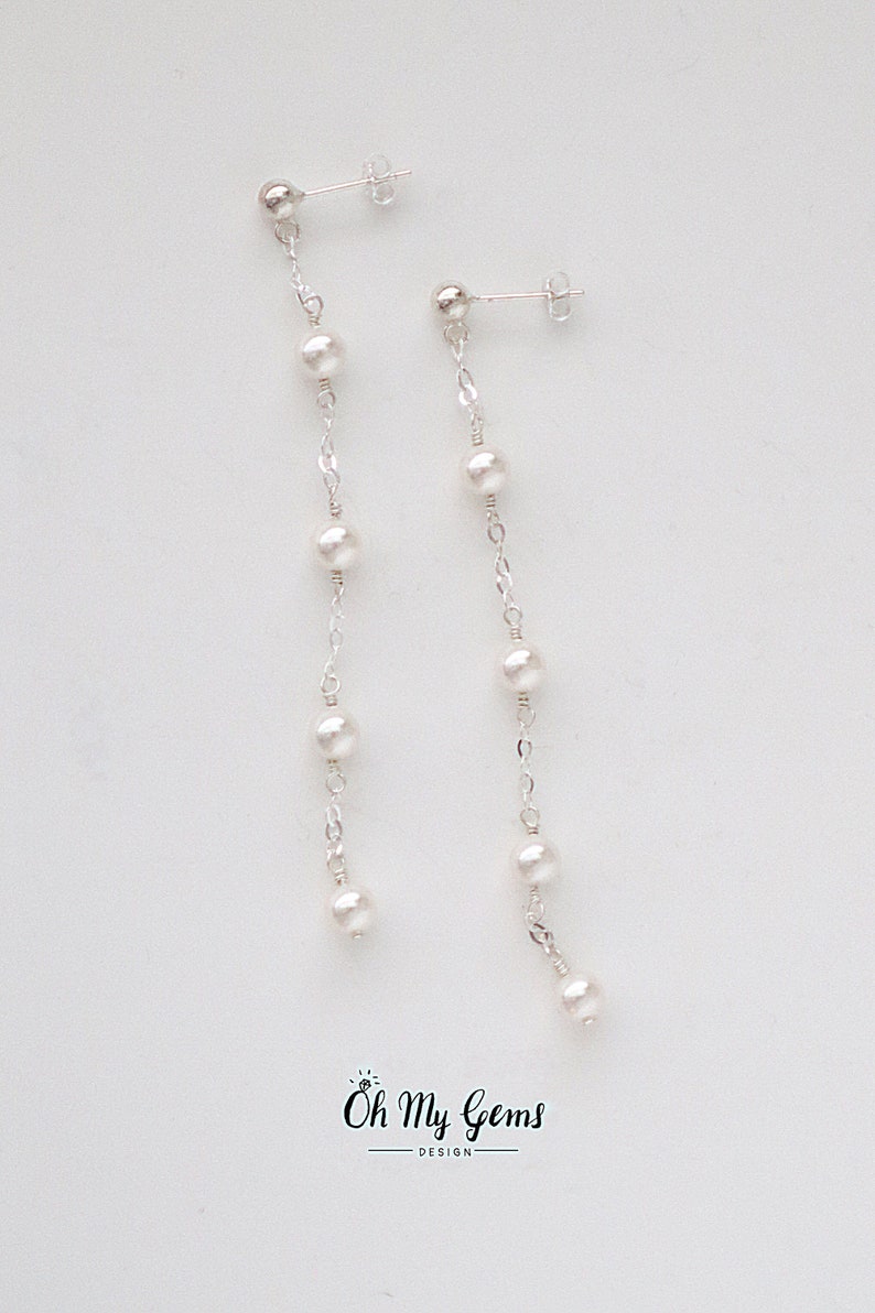 Puede incluir: Pendientes colgantes de plata con una serie de perlas blancas. Los pendientes tienen un cierre de poste y un dise&ntilde;o de cadena delicada. Los pendientes se muestran sobre un fondo blanco. El logotipo "Oh My Gems Design" est&aacute; en la parte inferior.