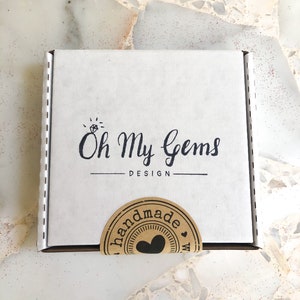 May include: A square white cardboard shipping box featuring the black printed text Oh My Gems DESIGN with a small diamond icon above the text. A circular brown paper sticker with the word handmade and a heart icon is sealed over the bottom edge.