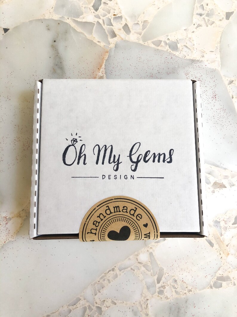 May include: A square white cardboard shipping box featuring the black printed text Oh My Gems DESIGN with a small diamond icon above the text. A circular brown paper sticker with the word handmade and a heart icon is sealed over the bottom edge.