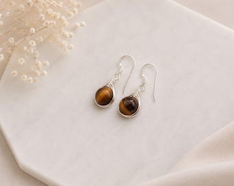 Puede incluir: Un par de pendientes colgantes con piedras preciosas de ojo de tigre redondas y pulidas. Cada piedra está envuelta en un diseño de alambre de tono plateado y suspendida de un gancho simple, creando un conjunto de joyería simétrico.