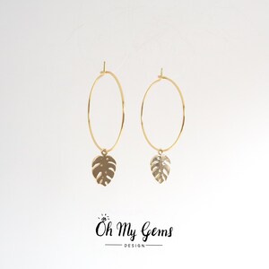 May include: A pair of gold hoop earrings featuring a dangling monstera leaf charm on each hoop. The text Oh My Gems DESIGN is printed in black at the bottom center of the image, with a small diamond icon above the letter O.