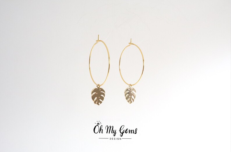 May include: A pair of gold hoop earrings featuring a dangling monstera leaf charm on each hoop. The text Oh My Gems DESIGN is printed in black at the bottom center of the image, with a small diamond icon above the letter O.
