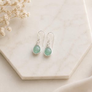 Peut inclure: Une paire de boucles d'oreilles pendantes avec des pierres vert clair lisses en forme de goutte. Chaque pierre est fixée par un fil métallique argenté enroulé sur le dessus et suspendue à un crochet d'oreille argenté simple et équilibré.