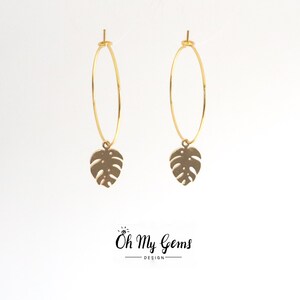 May include: A pair of gold-toned hoop earrings featuring a dangling monstera leaf charm. The earrings are displayed against a plain white background with the text Oh My Gems DESIGN printed in black at the bottom center of the image.
