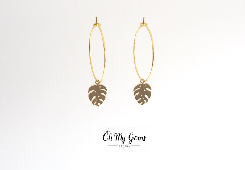May include: A pair of gold-toned hoop earrings featuring a dangling monstera leaf charm. The earrings are displayed against a plain white background with the text Oh My Gems DESIGN printed in black at the bottom center of the image.